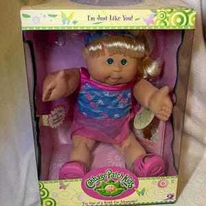 May include: Cabbage Patch Kids doll in a box. The doll has blonde hair, green eyes, and is wearing a pink and blue outfit with matching shoes. The box reads "I'm Just Like You!" and "I'm One of a Kind, I'm Adoptable!"