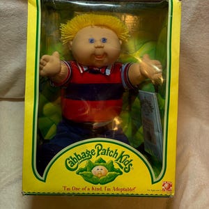 May include: A Cabbage Patch Kids doll with yellow yarn hair and blue eyes, wearing a red and navy blue striped shirt with a collar. The doll is inside a yellow cardboard box with the text Cabbage Patch Kids and I'm One of a Kind, I'm Adoptable.