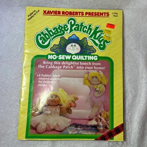 May include: A yellow Cabbage Patch Kids No-Sew Quilting pattern booklet by Xavier Roberts. The cover features green text, a central illustration of two blonde dolls in ballet outfits, and the title text: Bring this delightful bunch from The Cabbage Patch into your home! 8 Padded, fabric covered projects for children's rooms.