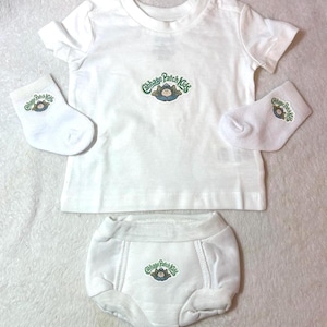 May include: A white short-sleeved t-shirt, a pair of white bloomers, and two white socks. Each item features a small green and brown Cabbage Patch Kids logo with a graphic of a doll emerging from a cabbage leaf centered on the fabric.