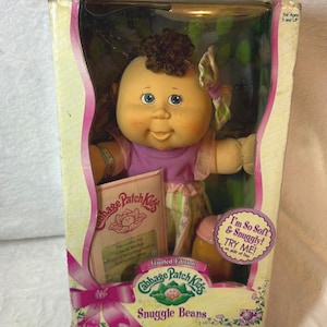 May include: A Cabbage Patch Kids Snuggle Beans doll in its original packaging. The doll has a soft, round face, blue eyes, and brown hair. It wears a purple top and patterned pants. The box includes text that reads "I'm So Soft & Snuggly! TRY ME!"