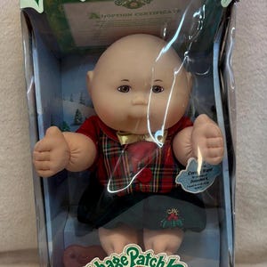 May include: A Cabbage Patch Kids Holiday Baby doll in a box. The doll has a bald head, light skin, and is wearing a red and green plaid vest over a dark green skirt. The packaging features holly leaves, the text Cabbage Patch Kids, and Holiday Baby.