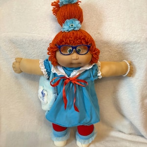 Può includere: Una bambola con capelli in lana arancione brillante raccolti in una coda alta con due accessori per capelli a forma di fiore azzurro. La bambola indossa occhiali blu, un vestito blu con colletto bianco e nastro rosso, leggings rossi e un bracciale.