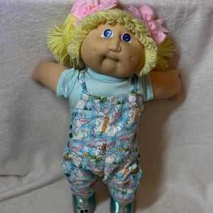 May include: A doll with yellow yarn hair styled in pigtails with pink bows, blue eyes, and dimpled cheeks. The doll wears a light blue short-sleeved shirt under blue and white checkered overalls featuring a print of white rabbits and carrots.