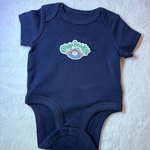 May include: A navy blue short-sleeved bodysuit featuring a Cabbage Patch Kids logo graphic on the chest. The garment has a round neckline, ribbed trim around the collar and leg openings, and three snap fasteners at the bottom crotch area.