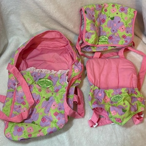 May include: A Cabbage Patch Kids doll carrier set featuring a lime green fabric with a purple and pink floral pattern. The set includes a soft pink-lined carrier with a white ruffled trim and a matching smaller harness with pink straps.