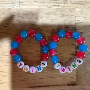 Puede incluir: Dos pulseras de cuentas circulares hechas de cuentas esféricas translúcidas rojas y azules alternadas. Cada pulsera presenta cuentas redondas blancas con letras negras. Una pulsera deletrea TWIN 1 y la otra TWIN 2 con un número 2 verde.