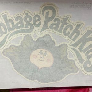May include: A rectangular white sticker featuring the Cabbage Patch Kids logo in a stylized grey font. Below the text is a grey illustration of a cabbage leaf surrounding a round, pale face with simple facial features and rosy cheeks.