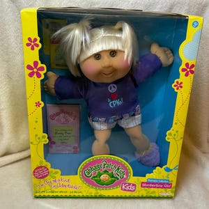 Puede incluir: Una muñeca Cabbage Patch Kids Slumbertime Girl con cabello rubio en coletas, vistiendo una camiseta morada de manga larga con un símbolo de paz, un corazón rojo y el texto "& CPK!" impreso al frente, junto a pantalones cortos claros estampados.