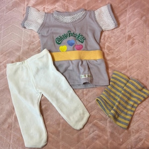 May include: A doll outfit consisting of a light gray short-sleeved shirt with white mesh sleeves, a yellow waistband, and the text Cabbage Patch Kids with three colorful hearts. It includes plain white leggings and a pair of yellow and gray striped leg warmers.