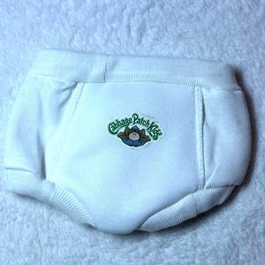 May include: White fabric nappy cover featuring an elastic waistband and leg openings. The centre of the garment displays a circular embroidered patch with the green text Cabbage Patch Kids above a small illustration of a doll character.