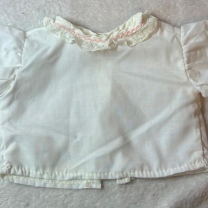May include: A white short-sleeved blouse featuring a ruffled collar and sleeve cuffs trimmed with delicate white lace and a thin pink ribbon detail. The garment has a button-down front closure and a simple, clean design with puffed sleeves.