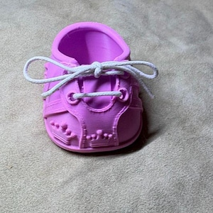 May include: A bright pink doll shoe featuring a moulded crown pattern on the sides and front. The shoe has a rounded toe, a high ankle opening, and is laced with a thin white cord tied in a bow across the top of the tongue area.
