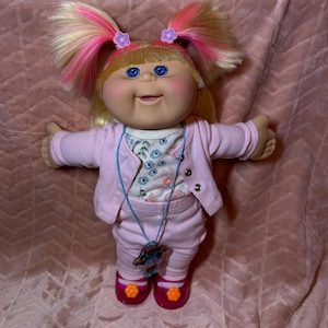 May include: A doll with blonde hair featuring pink highlights styled in two high pigtails with purple flower clips. The doll has blue eyes, wears a light pink cardigan over a floral print top, matching pink trousers, and pink shoes with orange flower accents.
