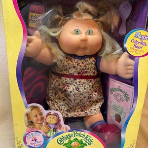 May include: A Cabbage Patch Kids Magic Touch ColorSilk doll with blonde hair in pigtails, green eyes, and a floral print dress. The doll is packaged in a yellow and purple box featuring the text Magic Touch ColorSilk Kids and I'm Adoptable.