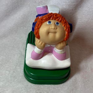 May include: A Cabbage Patch Kids figurine featuring a character with orange yarn-like hair, blue eyes, and a tan complexion. The figure wears a pink and white outfit, rests on a white pillow, and is mounted on a dark green rectangular base.