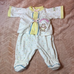 Op de afbeelding: Een wit baby-outfit met lange mouwen en een patroon van kleine veelkleurige hartjes. De top heeft een gele kraag, twee roze lintstrikjes en een geborduurde teddybeer met de tekst Cabbage Patch Kids. De broek heeft gesloten voetjes.