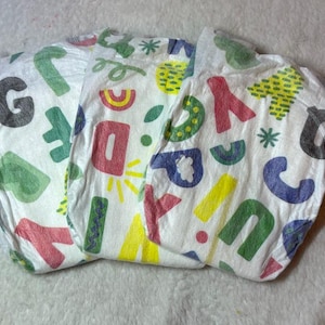 May include: A white disposable diaper featuring a colorful all-over print of various letters of the alphabet in red, green, blue, yellow, and black, along with small graphic shapes like stars and sunbursts scattered across the absorbent material.