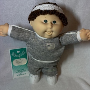 May include: A Cabbage Patch Kids doll wearing a gray sweatsuit with white trim and a white cat patch. The doll has brown curly hair, a white headband, and green eyes. The doll is wearing white sneakers and has a birth certificate.