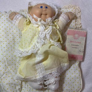 May include: A Cabbage Patch Kids doll with a round vinyl head, blue eyes, and a dimpled smile. The doll wears a pale yellow dress with white lace trim, a matching bonnet, and white booties. A birth certificate labelled Ruth Hedda is included.