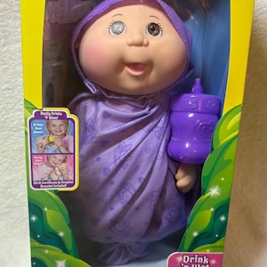 Op de afbeelding: Cabbage Patch Kids "Drink 'n Wet" pasgeboren pop in een doos. De pop draagt een paarse outfit en houdt een paarse fles vast. De doos heeft een gele rand en groene bladaccenten. Op de doos staat "Really Drinks Wets!" en "Birth Certificate & Hospital Bracelet Included!"