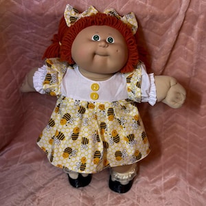 Pode incluir: Uma boneca com cabelo de lã ruivo em maria-chiquinhas com laços a combinar, olhos verdes e rosto com covinhas. A boneca usa um corpete branco com dois botões amarelos e uma saia amarela com um padrão repetido de abelhas e favos de mel.