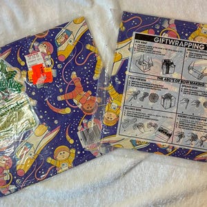 May include: Two sheets of vintage wrapping paper featuring a space theme with astronauts, rockets, and planets on a dark blue background. One sheet is still in its original packaging and includes a price tag. The other sheet has gift wrapping tips.