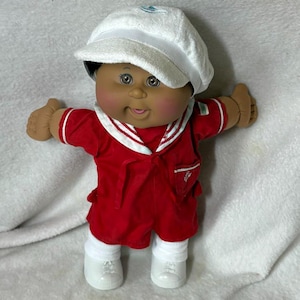May include: A doll with a round face, dark skin, and brown eyes wearing a white fabric cap, a red sailor-style shirt with a white collar, white socks, and white shoes. The doll has a small embroidered detail on the pocket of the red shirt.