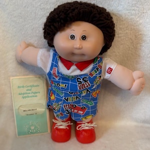 May include: A doll with dark brown yarn hair, brown eyes, and a soft vinyl face. It wears a white shirt with a red collar under blue overalls featuring a colorful traffic-themed print. Red shoes are on its feet. Beside it is a birth certificate.