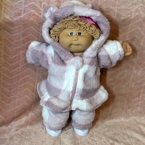 May include: A doll with tan skin, green eyes, and curly blonde yarn hair wearing a fuzzy, hooded, zip-up jumpsuit in a white and dusty purple plaid pattern. The hood features small ears, and a bright pink bow is tucked into the hair.