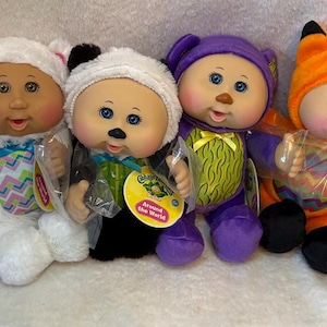 May include: Four Cabbage Patch Kids dolls dressed in animal-themed costumes. The dolls feature plush outfits resembling a white bear, a black and white panda, a purple bear, and an orange fox. Each doll has a round face, rosy cheeks, and a tag.