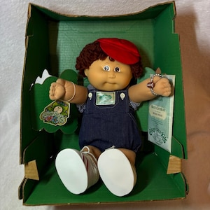 Pode incluir: Uma boneca Cabbage Patch Kids com cabelo de lã castanho, boné vermelho e jardineiras de ganga azul-escura sobre uma camisa aos quadrados azuis e brancos. A boneca usa sapatos brancos e está numa caixa de cartão verde com certificado.