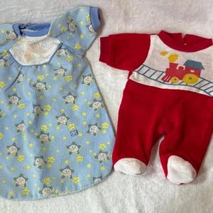 May include: Two baby outfits. One is a light blue sleep sack with a pattern of stars and smiling faces. The other is a red onesie with a train graphic on the chest. Both are made of soft fabric.