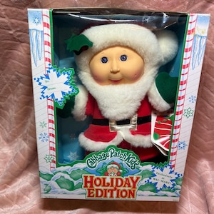May include: A Cabbage Patch Kids Holiday Edition doll dressed as Santa Claus with a white faux fur-trimmed red suit, green mittens, and a matching hat. The doll is inside a rectangular cardboard box featuring candy cane borders and text.