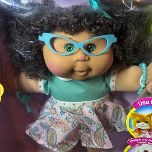 Könnte beinhalten: Eine Cabbage Patch Kids Puppe mit dunklem, lockigem Haar, grünen Augen und hellblauer Katzenaugenbrille. Die Puppe trägt ein türkisfarbenes Oberteil mit orangefarbenen Punkten und einen rosa Rock mit Paisleymuster, ein blaues Perlenarmband und blaue Schuhe.
