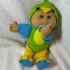 Puede incluir: Una muñeca Cabbage Patch Kids con un disfraz de pájaro verde con pico amarillo y ojos negros. La muñeca tiene un chupete azul en la boca y lleva ropa azul y amarilla.
