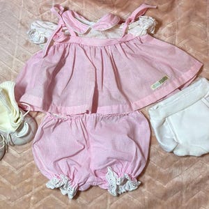 May include: A doll clothing set featuring a pink and white gingham top with a Peter Pan collar and tie straps, matching pink gingham bloomers with white eyelet lace trim, a pair of white shoes with socks, and a pair of plain white diaper-style briefs.