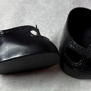 May include: A pair of small, black doll shoes. The shoes have a glossy finish, a T-strap design, and decorative stitching around the edges. The soles are solid black.