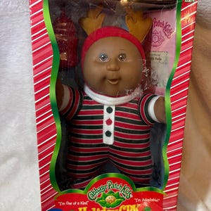 Pode incluir: Uma boneca Cabbage Patch Kids com um pijama de riscas vermelhas e verdes e um gorro vermelho a condizer com hastes de rena castanho-claras. A boneca está numa caixa retangular com bordas de riscas vermelhas e brancas e o texto Holiday CPK.
