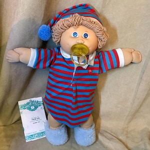 May include: A Cabbage Patch Kids doll with yarn hair, blue eyes, and a yellow dummy. The doll wears a red and blue striped shirt and matching nightcap with a blue pom-pom, plus light blue booties. A small white card with text is included.
