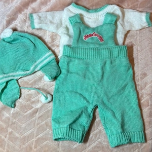 May include: A knitted doll outfit consisting of a white long-sleeve sweater with mint green trim, mint green overalls with the red embroidered text Cabbage Patch Kids on the bib, and a matching mint green knit hat with white stripes and pom-poms.