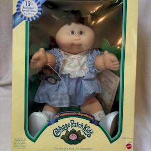 May include: A Cabbage Patch Kids 15th Anniversary Special Edition doll in its original packaging. The doll has brown yarn hair, brown eyes, and wears a blue and white gingham dress with a white lace-trimmed collar. The box includes text.