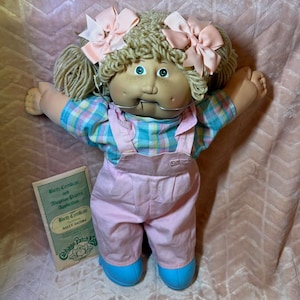 May include: A Cabbage Patch Kids doll with blonde yarn hair in pigtails, adorned with two light pink bows. The doll wears a plaid shirt in blue, pink, and white, paired with pink overalls and blue shoes. A birth certificate document is included.
