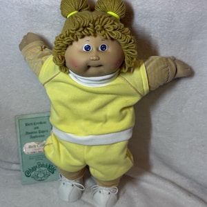 May include: A Cabbage Patch Kids doll with blonde yarn hair styled in two top knots. The doll wears a yellow outfit with a white turtleneck and white shoes. A birth certificate is visible.