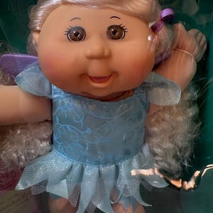 以下が含まれることがあります： 妖精の衣装を着たキャベツ人形。人形は金髪、茶色の目、バラ色の頬をしています。キラキラした多層スカートと紫色のアクセントが付いた水色のドレスを着ています。人形は紫色の妖精の羽を持っています。