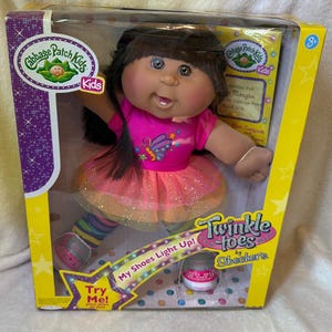 May include: A Cabbage Patch Kids doll with dark brown hair in a pink shirt featuring a butterfly graphic, a glittery orange and pink tutu, and multi-colored striped leggings. The doll is in a yellow and purple box labeled Twinkle toes by Skechers.