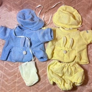 May include: A light blue terry cloth shirt with a bunny pocket and matching hat, a yellow terry cloth shirt with a bunny pocket and matching hat, a pair of yellow terry cloth bloomers, and a single light yellow sock, all made of soft fabric.