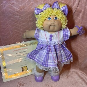 May include: A doll with yellow yarn hair styled in two pigtails with purple plaid bows, blue eyes, and dimples. It wears a white and purple plaid dress with lace trim, purple buttons, and a matching purple butterfly wrist accessory, accompanied by a certificate.