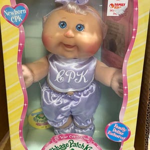 Pode incluir: Uma boneca Cabbage Patch Kids Newborn numa caixa. A boneca tem pele clara, olhos azuis e um traje lavanda com "CPK" no babete. A caixa tem o texto "25 Year Celebration" e "Family Dollar Exclusive!"