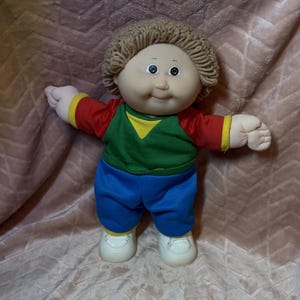 May include: A doll with light skin, brown yarn hair, and dark eyes. The doll wears a red shirt with yellow trim, a green chest panel, blue pants, and white shoes. The doll has a round face, a small nose, and a subtle smile, with arms extended outward.